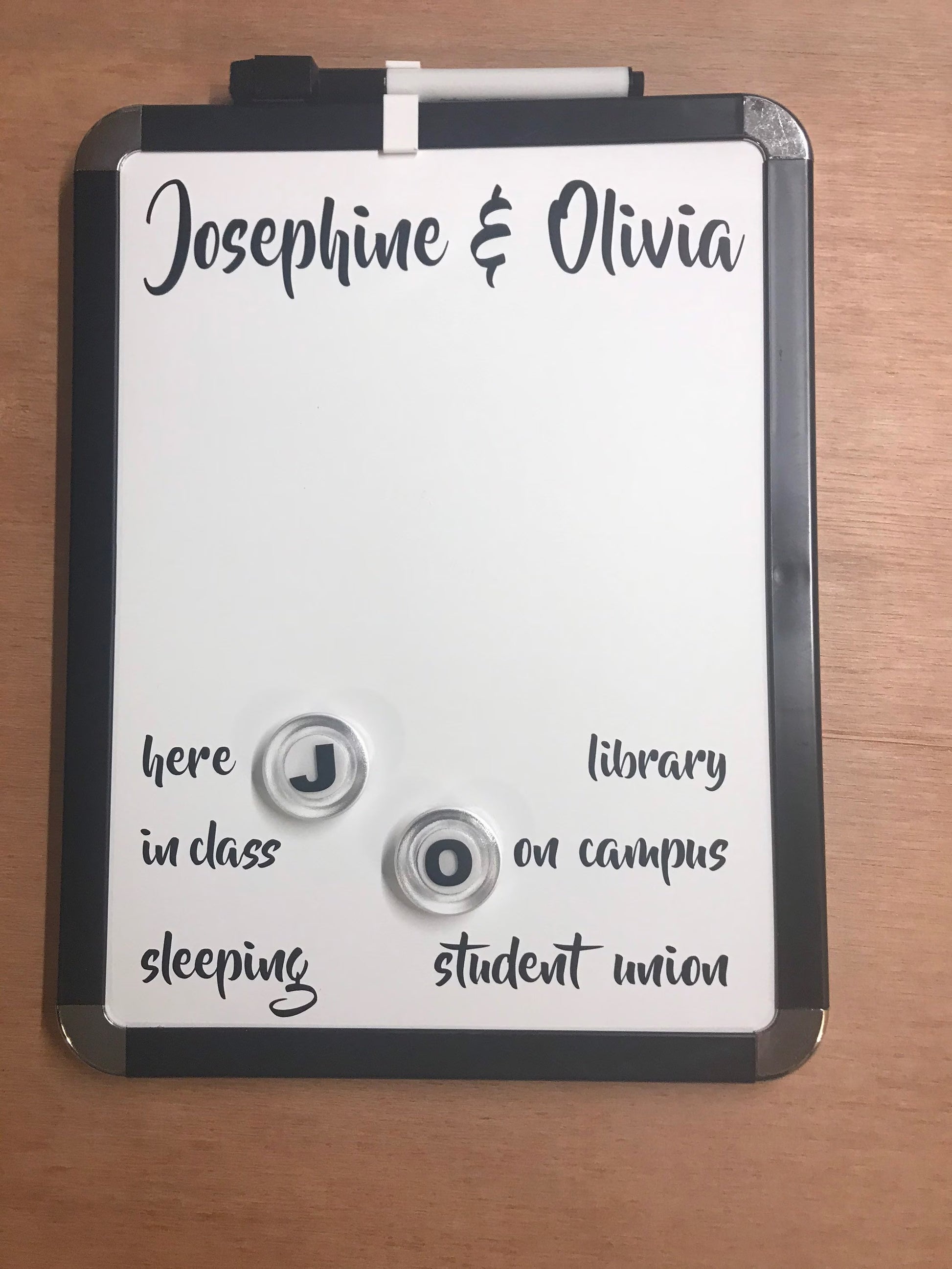 A personalized dry-erase whiteboard with a black frame and two silver magnetic letters, shown on a wooden tabletop. The whiteboard is customized with the names 'Josephine & Olivia' at the top and word associations for the letters 'J' and 'O' below.