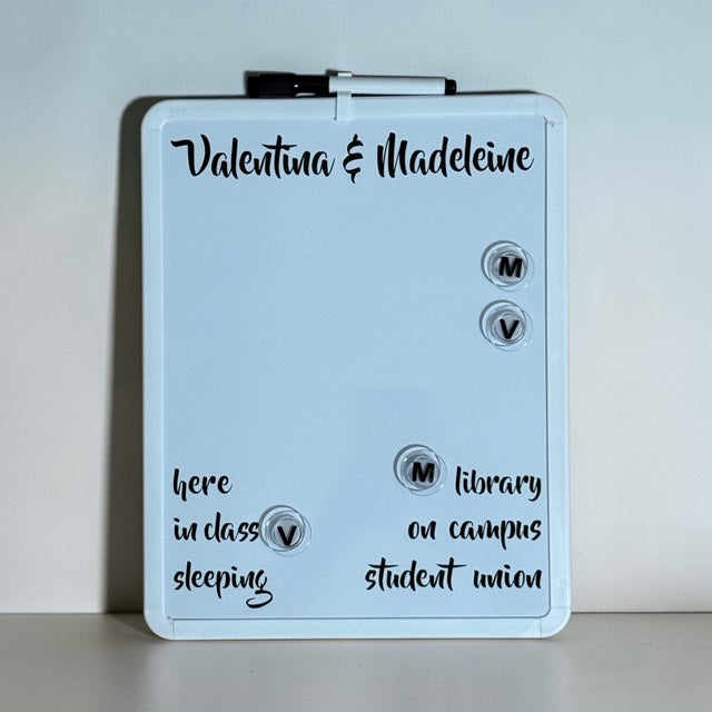 A personalized white dry-erase board with a black marker clipped at the top, featuring custom names and handwritten notes on a light gray background.