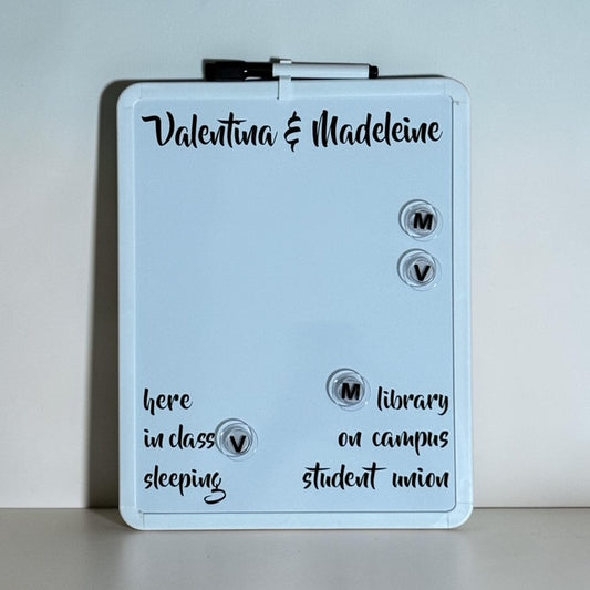 A personalized white dry-erase board with a black marker clipped at the top, featuring custom names and handwritten notes on a light gray background.
