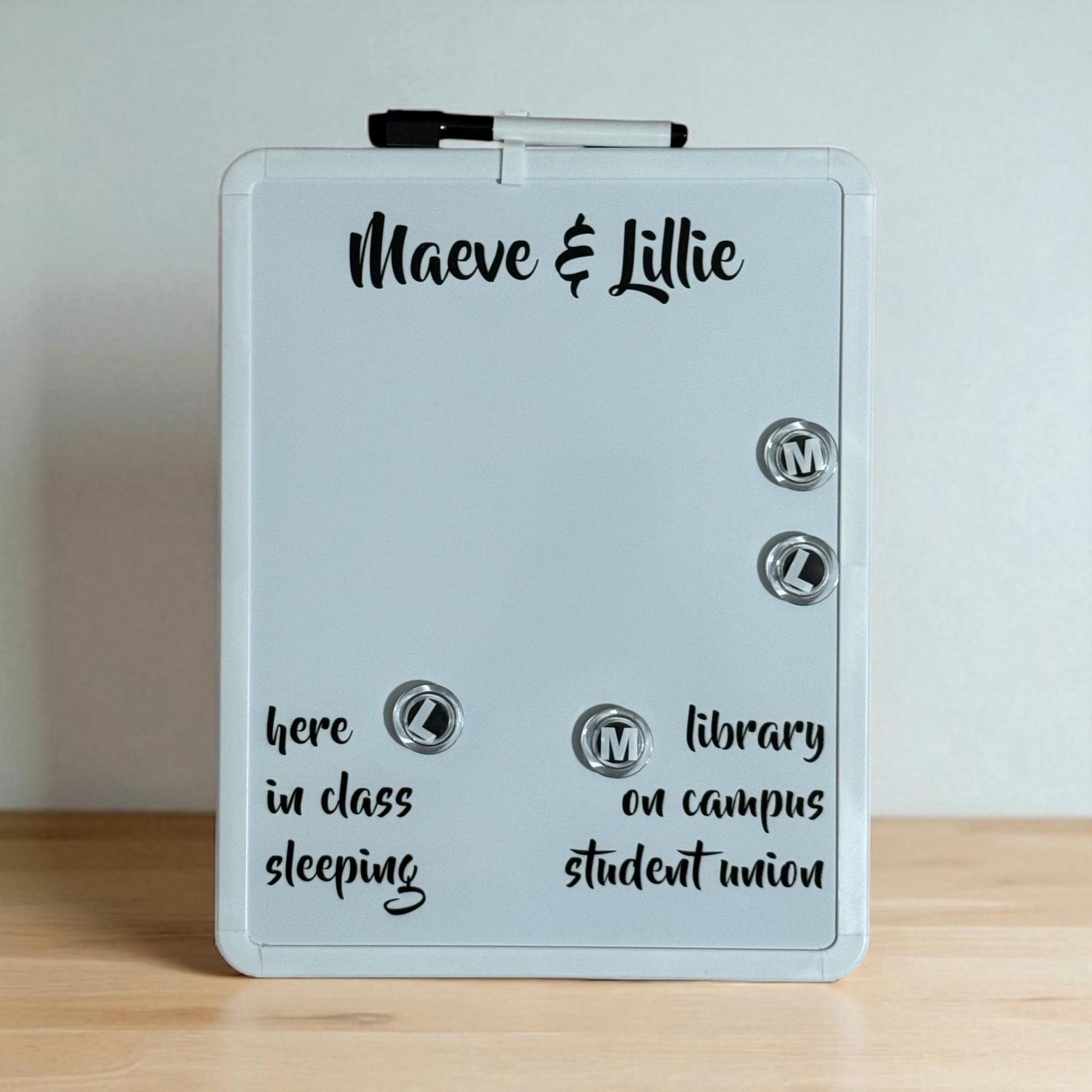 A white magnetic dry-erase board with a black marker on top, personalized with the names 'Maeve & Lillie' and sample text, standing on a light wood surface against a neutral light gray wall.