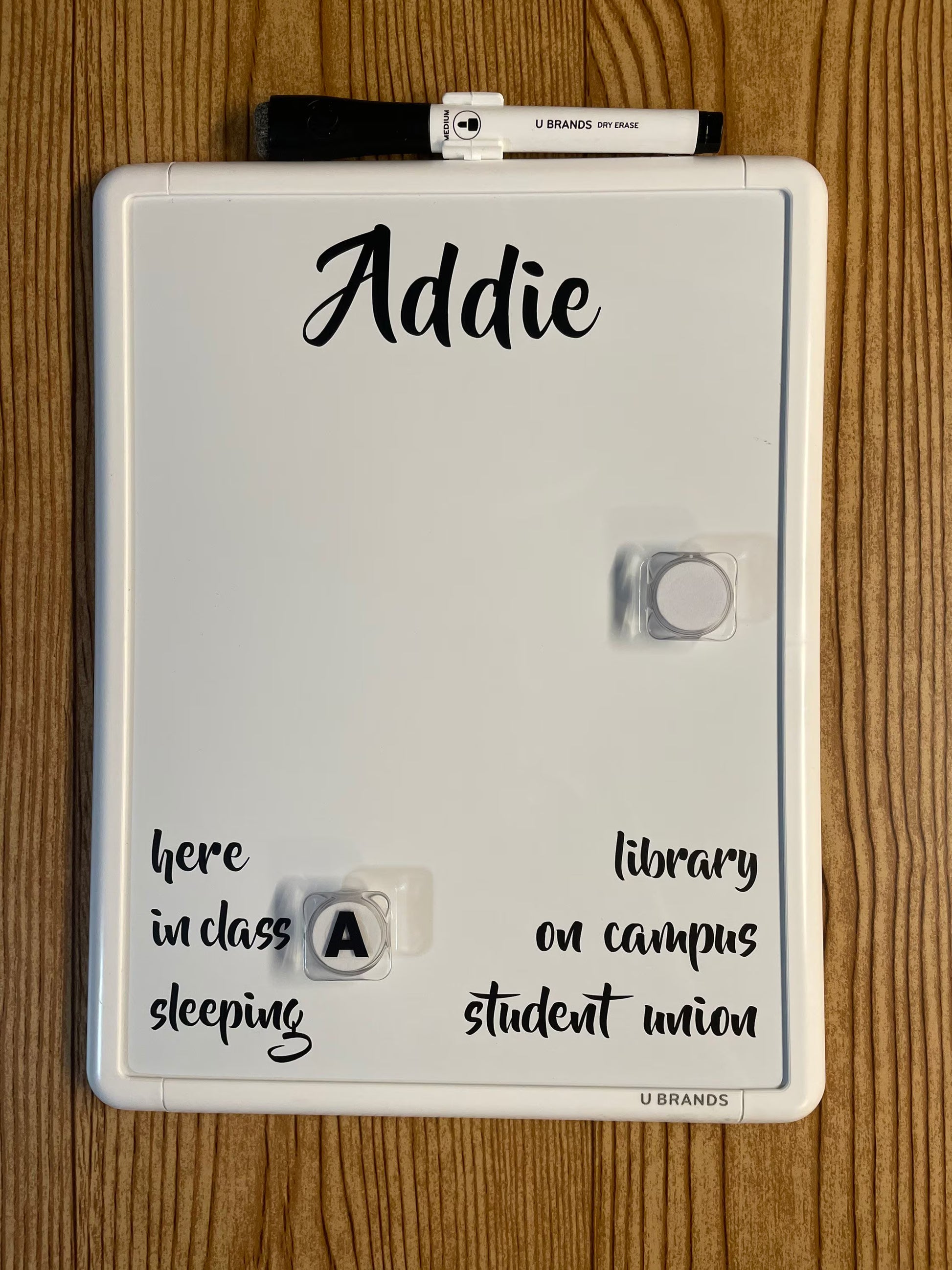 A white magnetic dry-erase board with a black marker on top, personalized with the name 'Addie' in black script. The board is photographed flat on a light brown wood-grain surface.